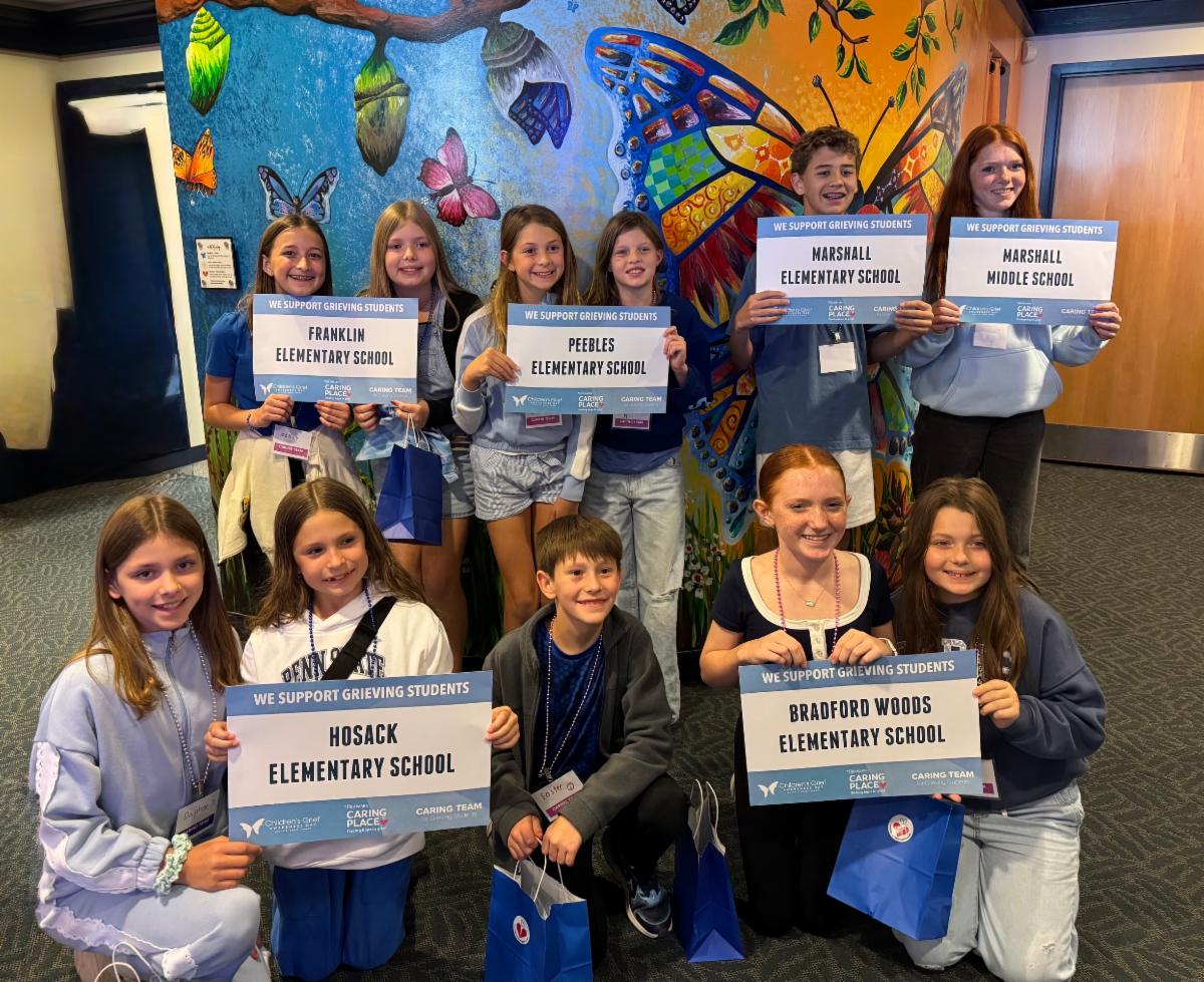 A group of North Allegheny student leaders poses together in front of a colorful butterfly mural at the Highmark Caring Place. The students hold signs reading "We Support Grieving Students" representing Franklin Elementary, Peebles Elementary, Marshall Elementary, Marshall Middle, Hosack Elementary, and Bradford Woods Elementary schools.