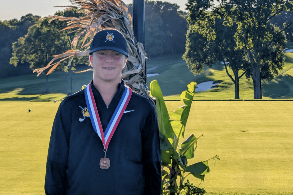 North Allegheny golfer Jake Hendrick stands outdoors on a sunlit golf course wearing a dark long-sleeve athletic shirt, a navy cap with a tiger paw emblem, and a red, white, and blue award medal around his neck. Rolling green fairways and trees are visible in the background under a clear sky.