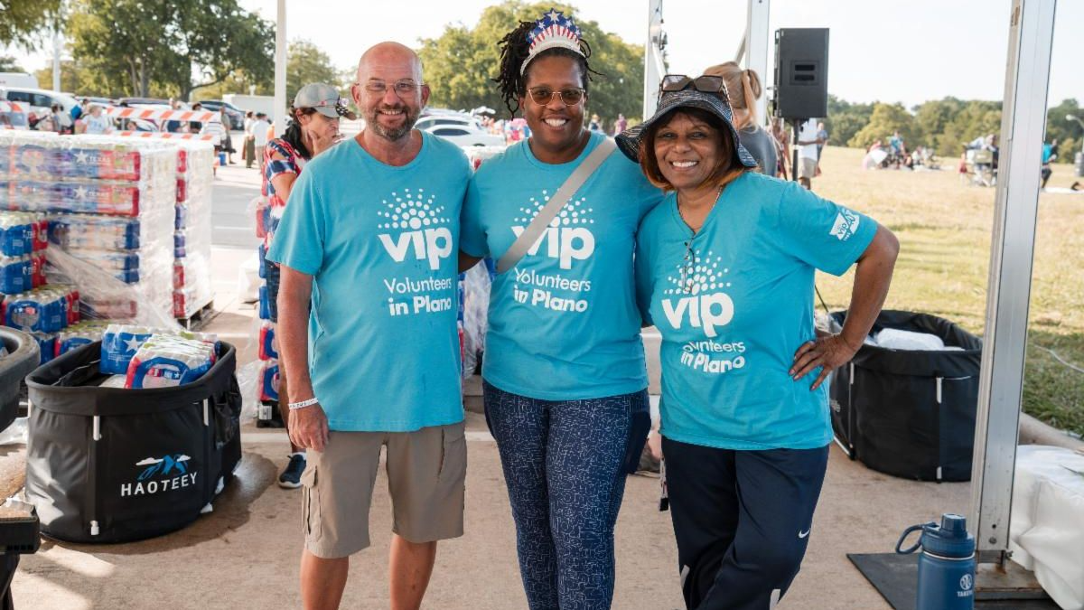 Three people in light blue VIP shirts smiling in front of pallet of water bottles