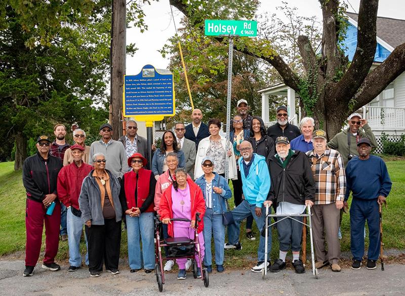 Photograph shows diverse community group posing outdoors beside green “Holsey Rd” street sign and blue historical marker. Adults and children, including several seniors and people using wheelchairs or walkers, gather before a house and trees for a commemorative community event.
