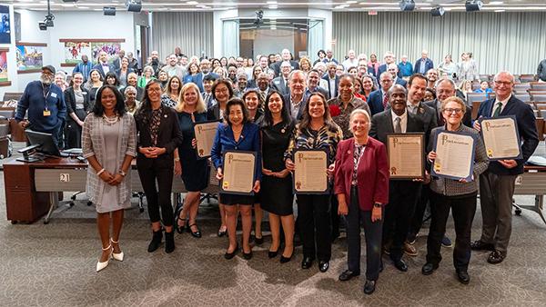 Group photograph shows a large, diverse gathering of professionals posing in a conference room. Several attendees hold framed certificates, indicating an awards or recognition ceremony, with tables, cameras, banners, and seated observers visible behind them.