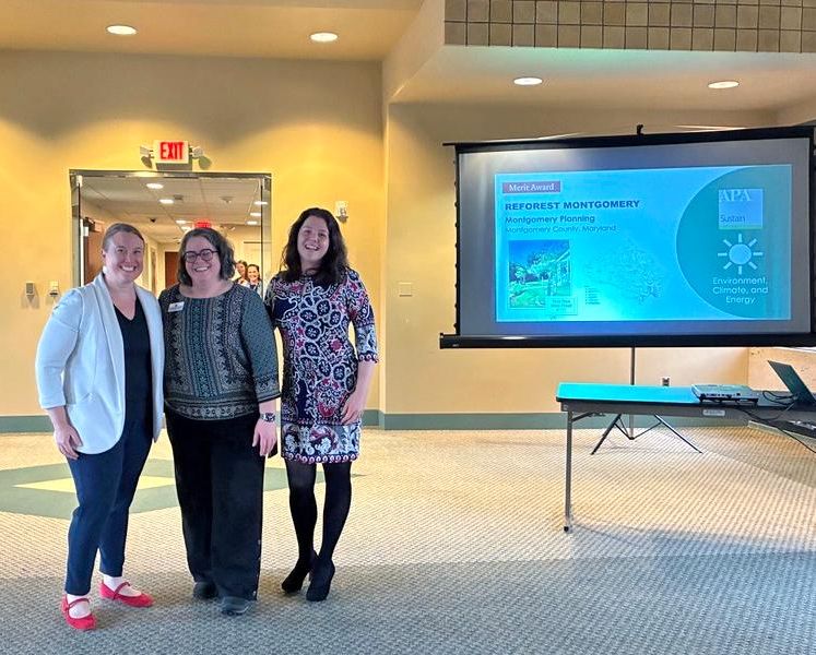 Three people standing indoors, smiling at the camera. A presentation slide behind them displays "Reforest Montgomery" and "Excellence in Climate and Energy" with logos from APA and National Capital Area Chapter. A table with a laptop is on the side. 