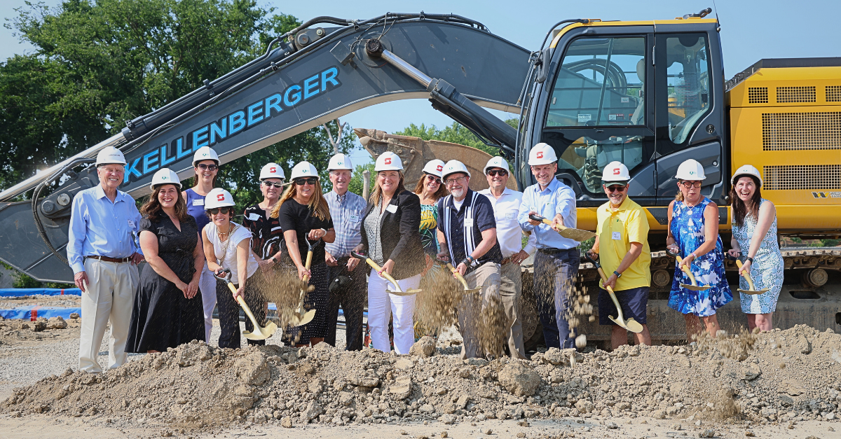 Professionally dressed people wear white hard hats and toss shovels of dirt at a construction site with a large digger in the background.