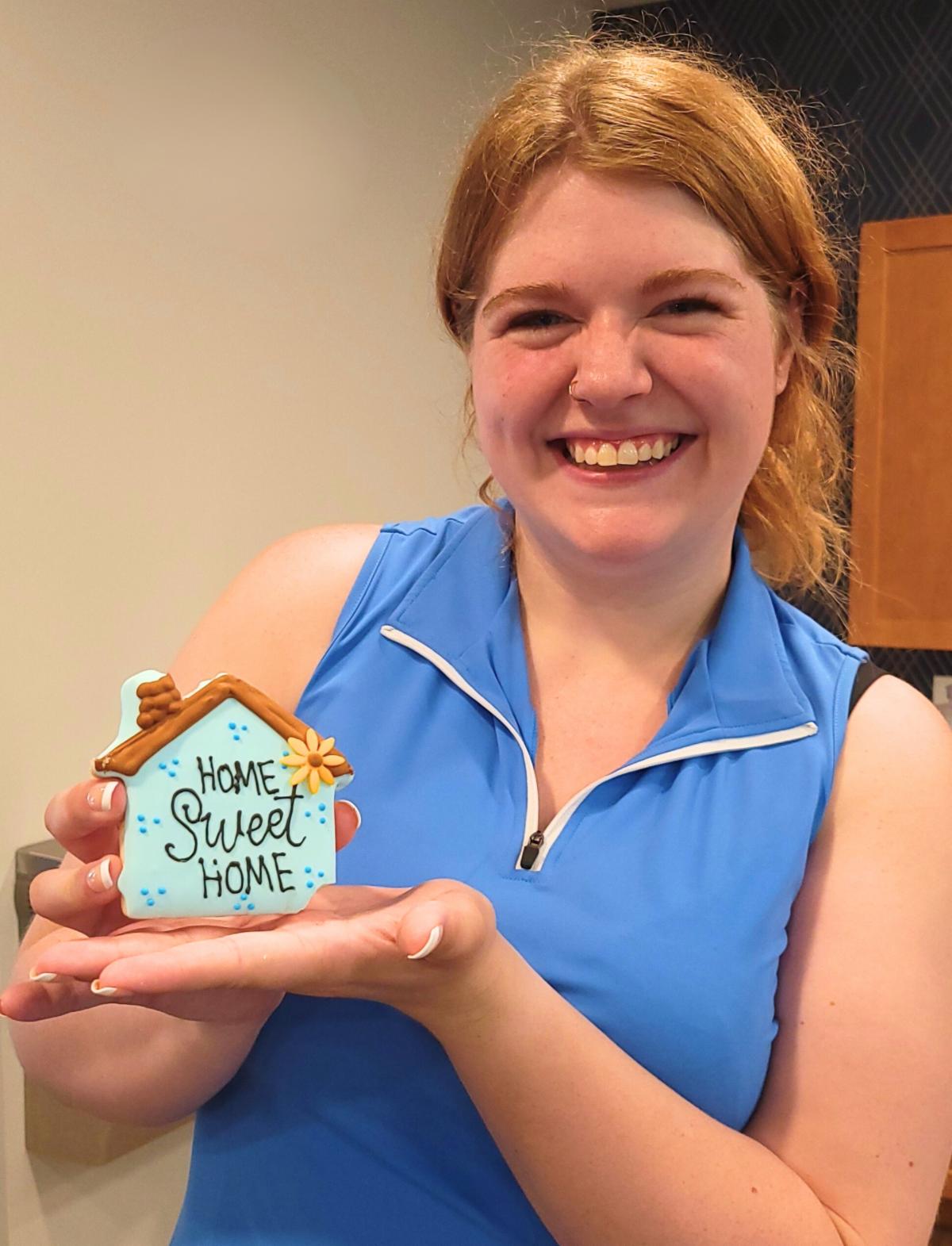 A young woman with reddish-blonde hair wearing a blue sleeveless shirt smiles as she poses with a house shaped cookie that says Home Sweet Home.