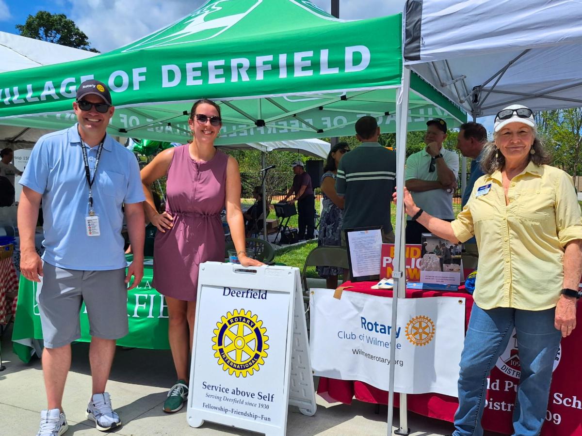 Three people pose under a green event tent that says Village of Deerfield at a summer event.
