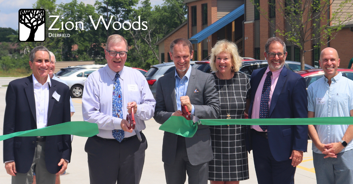 Professionally dressed people smile and pose with a large green ribbon that is being cut with large scissors in front of an apartment building.