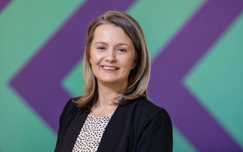 Person wearing a black blazer over a white top with a black dotted pattern standing in front of a background featuring large diagonal purple and green stripes. The setting appears indoors with bright even lighting.
