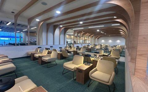 Modern airport lounge with rows of cream armchairs and small tables arranged in sections. curved wooden ceiling beams and soft lighting throughout a spacious seating area with large windows in the background.