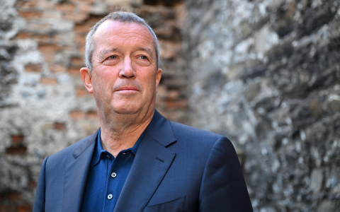 Person wearing a dark blue blazer and navy shirt standing in front of a textured stone and brick wall with an aged architectural appearance