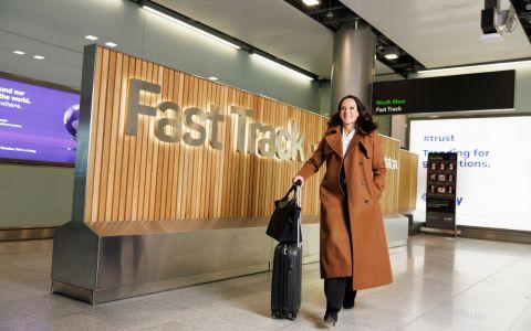 A person walks through an airport area pulling a small suitcase. A large sign reading Fast Track is displayed on a wooden panel behind them.