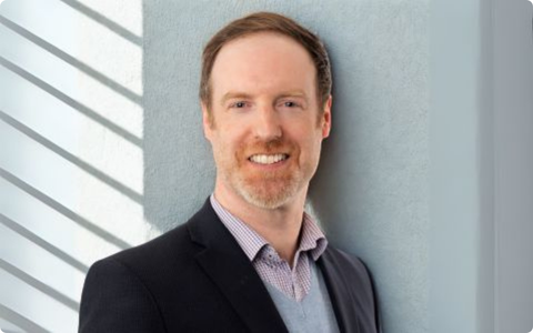 The image shows a person wearing a dark blazer over a light blue sweater and a patterned collared shirt. The face of the person is blurred out. The background features a wall with diagonal shadows. There are no other visible elements or text in the image.