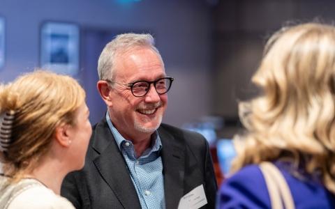 Photograph of three people in conversation at a networking event. One person wearing glasses and a dark blazer stands in the centre while two other attendees are shown from the side in the foreground. The background is softly blurred with indoor lighting.