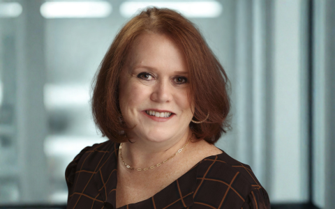 Professional headshot of a person with shoulder-length hair wearing a dark patterned top. The photograph is taken indoors against a softly blurred office background with natural light.