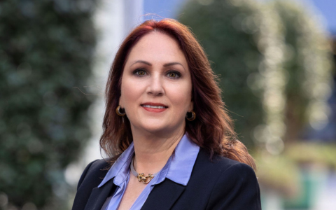 Person with long dark hair and light highlights wearing a dark blazer and light blue shirt standing outdoors with a blurred background of greenery and building