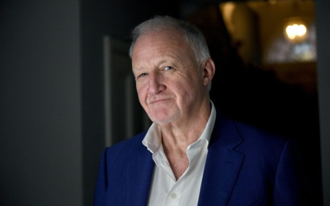 Professional portrait of an individual wearing a blue blazer and white shirt, standing indoors against a softly blurred background. The image is a head-and-shoulders photograph with natural lighting and a neutral backdrop.