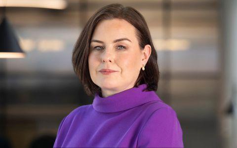 Professional headshot of a person wearing a purple high neck top against a softly blurred indoor background used for speaker or member profile.