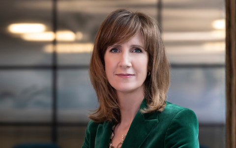 A person with shoulder‑length light brown hair wearing a deep green velvet blazer and a necklace, seated indoors in a modern office setting with frosted glass panels and warm overhead lighting in the background