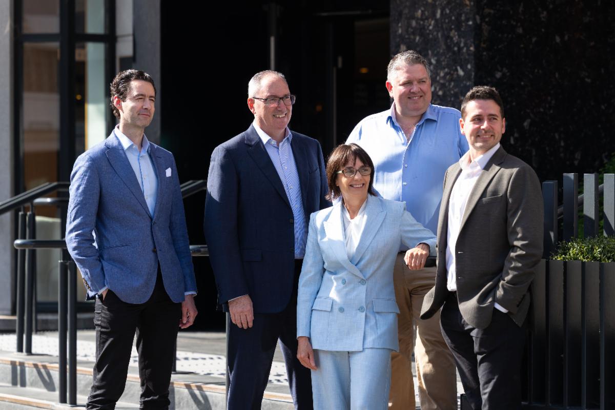 five people in business attire posing for a photograph outside a building in the sunshine