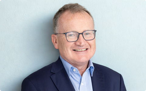 The image shows a person standing against a light background wearing a dark blue suit jacket over a light blue dress shirt. Face to camera smiling. There are no symbols text or additional elements in the image