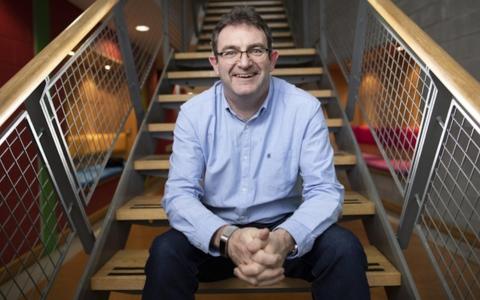 Person seated on a wooden stairs in a blue shirt and glasses. Smiling and looking to camera
