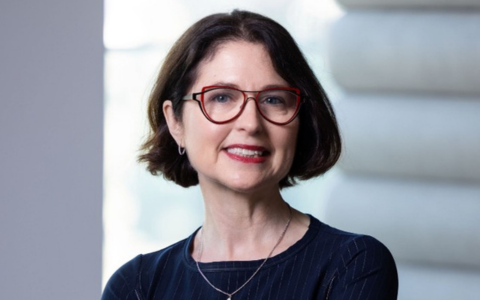 A woman stands indoors with her arms crossed wearing a long sleeved dark top and a delicate necklace. Soft natural light comes from a window on the left and a textured wall is visible in the background.