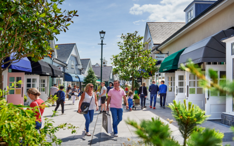 Outdoor shopping street with people walking and carrying shopping bags on a sunny day. The street is lined with boutique style shops featuring colourful awnings in shades of blue green and black. Trees and plants add greenery along the walkway. The sky is bright with scattered clouds creating a lively and vibrant atmosphere.