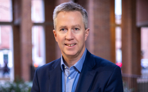 Person wearing a dark blue blazer over a light blue collared shirt standing in an outdoor setting with brick columns and large windows in the background. The lighting is natural and soft with hints of greenery and blurred urban elements visible behind the person.