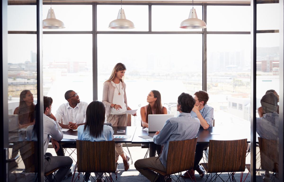 a group of people are sitting around a conference table in a modern office setting with large windows in the background one person is standing and appears to be leading the discussion while others are seated and listening attentively there are laptops and documents on the table