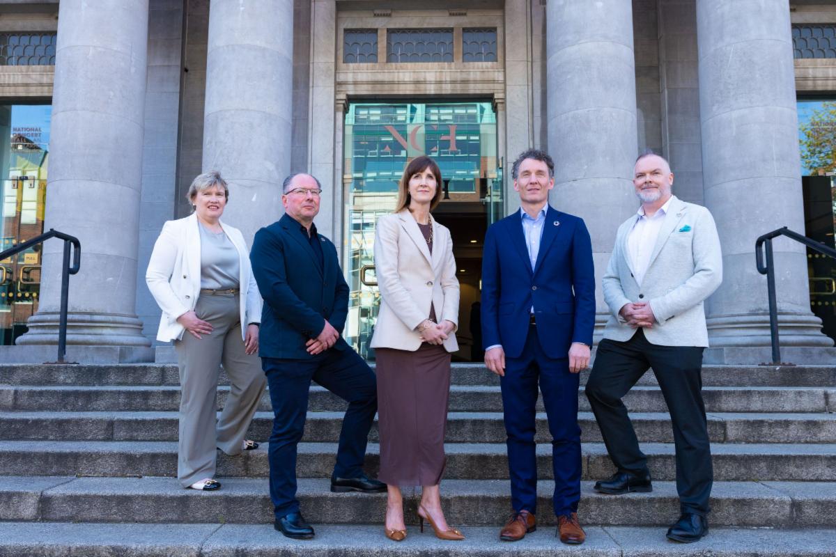Photograph of five professionals standing on the steps outside a large stone building with tall columns and glass doors. Group posed in business attire in front of the building entrance.