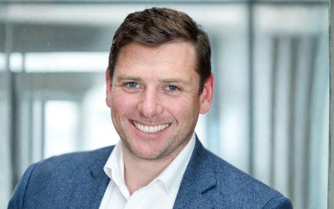 Professional headshot of a man wearing a blue jacket and white shirt standing in a modern office setting.