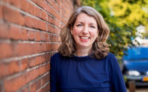 Professional portrait of a woman with shoulder length light brown hair wearing a dark blue top. She is standing outdoors beside a red brick wall with greenery and a parked car visible in the background.