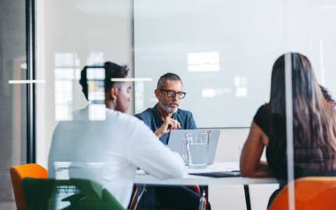 Group of people seated around a table in a modern office setting engaged in discussion. Laptops and documents are visible on the table. The scene is viewed through a glass partition creating reflections that add depth to the image. Bright natural light fills the room suggesting a collaborative work environment.