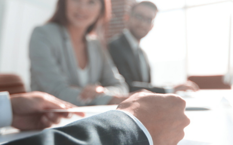 Close up of a business meeting setting with three people seated at a table wearing formal attire. The focus is on a hand holding a pen over documents in the foreground. Other participants are slightly out of focus in the background suggesting a professional discussion environment.