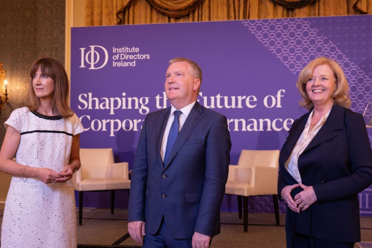 Three individuals stand in front of a purple backdrop displaying the text Institute of Directors Ireland and Shaping the future of Corporate Governance. A woman in a white dress is on the left a man in a blue suit and tie is in the center and a woman in a dark suit is on the right. Two empty chairs are positioned behind them