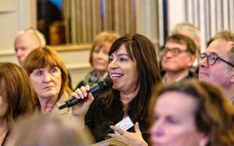 Two people seated in an audience during an indoor event wearing business attire. One person has shoulder length hair and a necklace while the other has slightly longer hair and a blazer. The background shows other attendees in soft focus with warm lighting creating a professional atmosphere.
