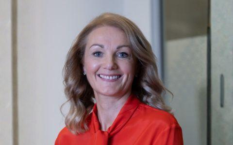 A person with shoulder length wavy hair stands indoors wearing a bright red blouse looks to camera