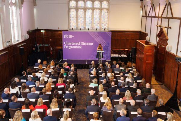 a large group of people is seated in an auditorium facing a stage where a woman is speaking at a podium with a purple backdrop that reads Chartered Director Programme