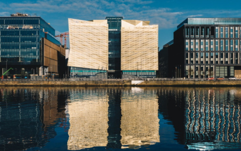 The image shows the Central Bank of Ireland. A modern architectural building with a unique design that includes a central structure made of angular golden colored panels The building is positioned between two other buildings that have more traditional glass and steel facades In front of the buildings there is calm water that reflects the entire scene creating a symmetrical visual effect.