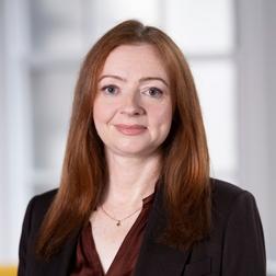 Professional headshot of an individual with long red hair, wearing a dark blazer and burgundy top, photographed against a softly blurred indoor background. The portrait is cropped from the shoulders up and features neutral professional attire suitable for a business or corporate profile.