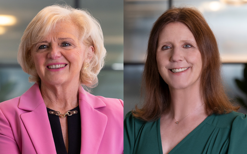 Two people side by side in an indoor setting with blurred office backgrounds one wearing a pink blazer and black top with light wavy hair and the other wearing a green top with long straight dark hair both smiling