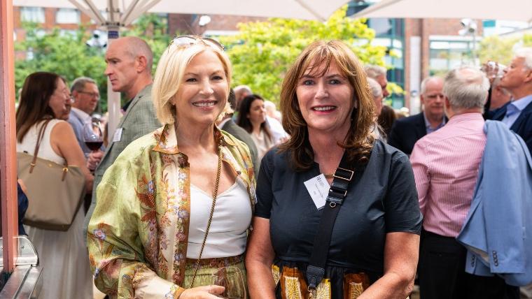 Two attendees standing together at an outdoor networking reception with other guests socialising in the background beneath large parasols.