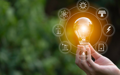 A hand holding a glowing light bulb outdoors, surrounded by circular energy‑related icons such as solar power, wind power, hydro, electricity, and sustainability symbols, with green foliage blurred in the background