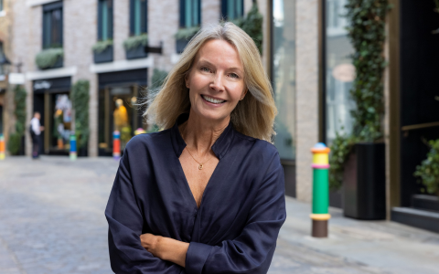Person standing on a cobblestone street in an urban setting wearing a dark long sleeve top with arms crossed. The background shows modern shopfronts with large windows and greenery on the walls along with colourful bollards lining the street. The scene is well lit suggesting daytime.