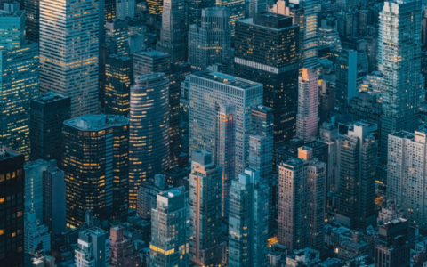 Aerial view of a dense cityscape featuring numerous high rise buildings and skyscrapers with varying architectural styles. The structures are closely packed creating a grid of streets barely visible between them. Many buildings have illuminated windows suggesting evening or early night time. The overall colour palette includes shades of blue grey and hints of warm light from interior lighting.