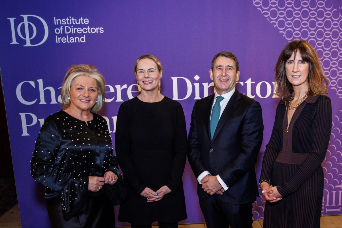 A group of four people stand in front of a purple Institute of Directors Ireland backdrop. They are dressed in formal attire and pose together for a photo.
