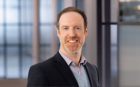 A person wearing a dark blazer over a light shirt and sweater stands in an indoor professional setting with soft blurred windows in the background.
