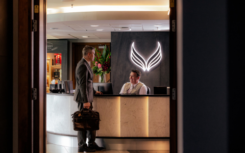 Photograph of a person standing at a hotel reception desk holding a briefcase. Another person is seated behind the desk. The reception area is viewed through an open doorway with a illuminated logo displayed on the wall behind the desk.