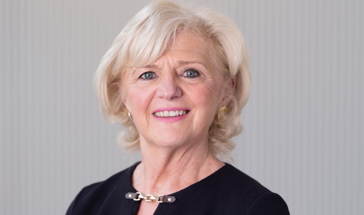 a person with short white hair wearing a black top with a chain detail at the neckline and gold earrings standing against a light grey striped 
