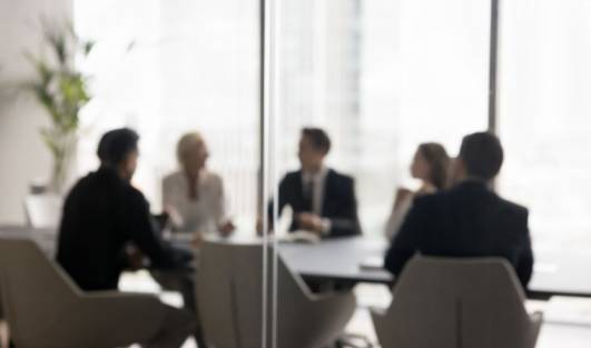 The image shows a group of five people sitting around a conference table in an office setting. The scene is viewed through a glass wall. The background includes large windows with an urban cityscape visible outside. There are no other visible elements or text in the image.