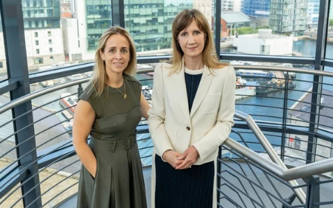Two individuals stand on an indoor balcony overlooking a waterfront cityscape with modern office buildings in the background. One individual wears a green dress and the other a light-coloured blazer over a dark outfit. Metal railings and dockside buildings are visible behind them.